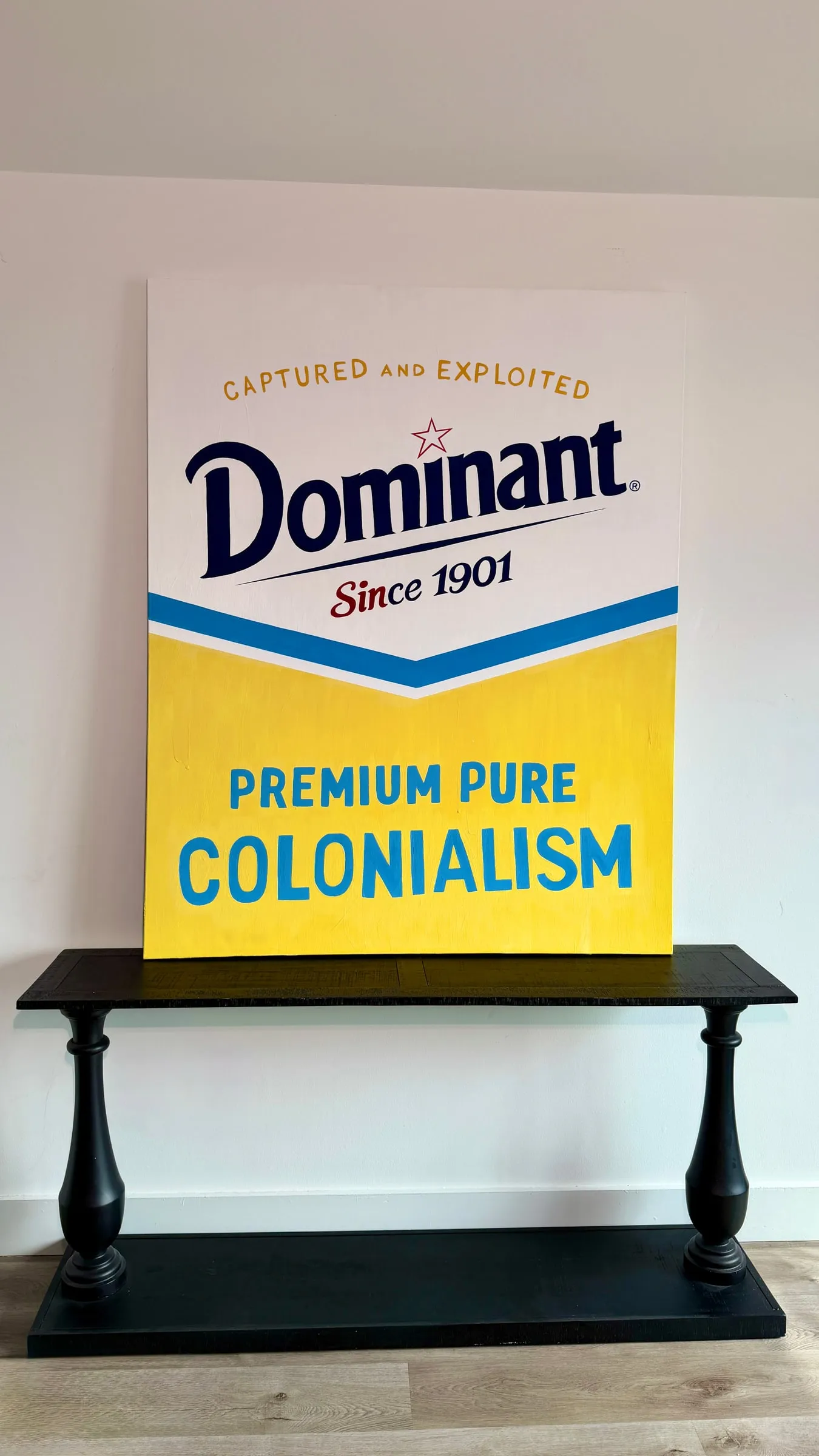 Painting of a yellow and white 'Dominant — Premium Pure Colonialism' sign, standing on a black console table