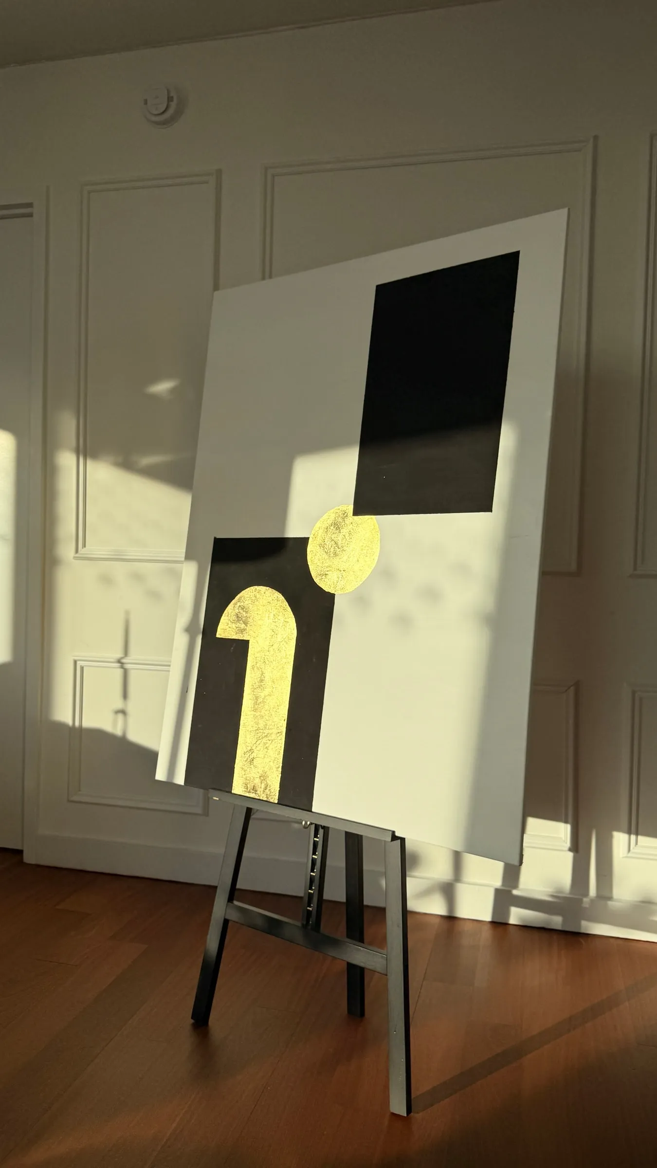 White canvas with a black rectangle and gold-leaf form, standing on an easel